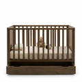 A dark brown wooden baby crib with vertical slats and a built-in storage drawer underneath, shown with a mattress, pillow, teddy bear, and small ball inside on a plain white background.