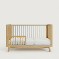 A light natural wood baby crib with vertical slats and angled legs, shown with a white mattress on a clean, solid off-white background.