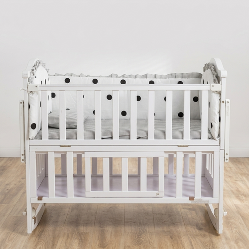 A white wooden baby crib with a lower storage shelf, styled with a gray and black polka dot bedding set, set against a plain white wall and light wood floor.