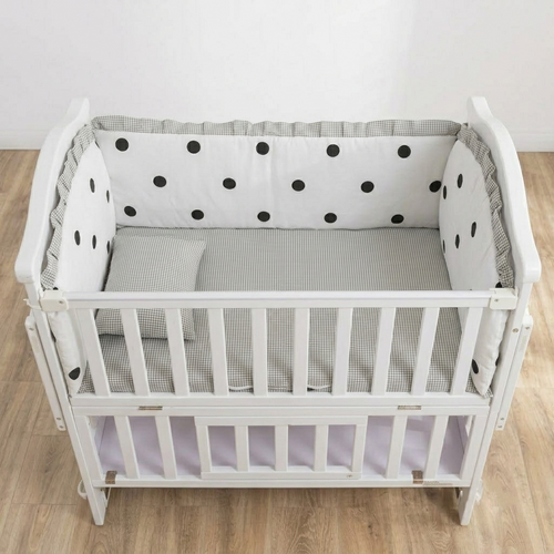 A white wooden baby crib with a gray and white gingham bedding set and white bumpers with black polka dots, shown on a light wood floor against a white wall.