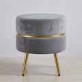 A round gray velvet ottoman stool with a gold metal band around the center and four tapered gold legs, shown on a light wood floor against a neutral gray background.