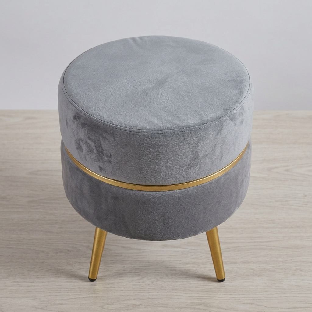 A round gray velvet stool with a gold metal band and three gold legs, set against a neutral light gray background and light wood floor.