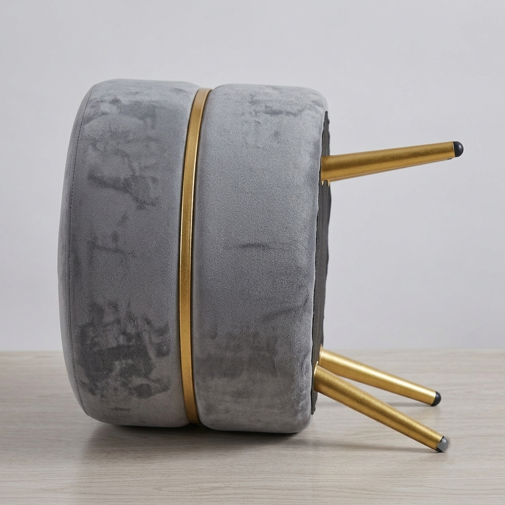 A round gray velvet stool shown upside down on a light wood surface, with gold metal legs and a gold vertical accent stripe.