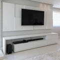 A modern white wooden media wall and matching low console in a bright living room, with a black flat-screen TV mounted above. The room has light wood flooring, white blinds, and a white rug.
