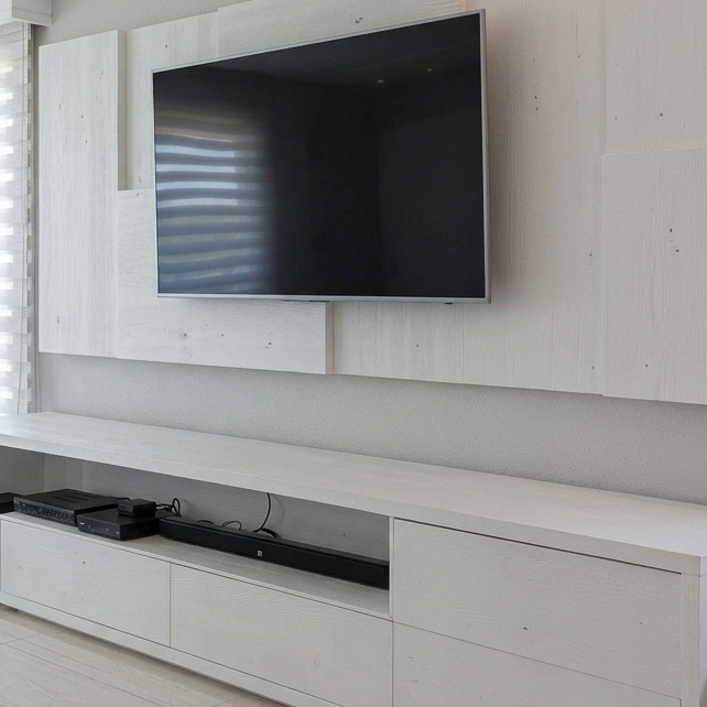 A modern wall-mounted flat-screen television with a slim silver bezel, displayed on a light wood floating media console and matching wall paneling.