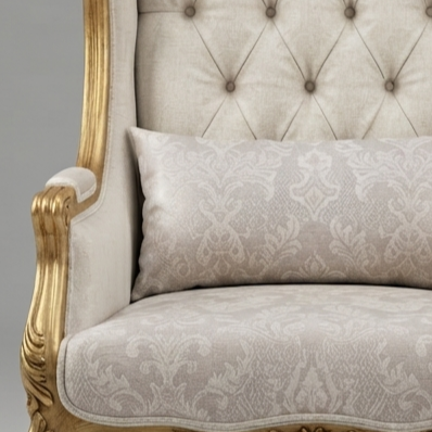 A close-up of an upholstered armchair with a matching lumbar pillow. The chair features a tufted cream fabric backrest and carved gold wooden arms, while the seat and pillow are covered in a light beige damask patterned fabric.