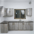 A compact L-shaped kitchenette with light gray wood-grain cabinets, dark gray countertops, a stainless steel sink, and gold-toned handles, set against a plain white wall.