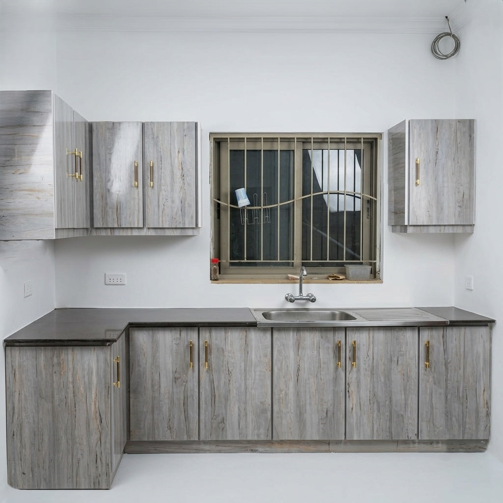 A compact L-shaped kitchenette with light gray wood-grain cabinets, dark gray countertops, a stainless steel sink, and gold-toned handles, set against a plain white wall.