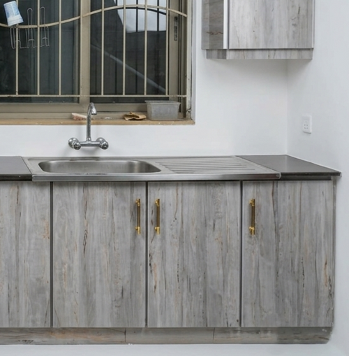 Gray wood-look kitchen cabinets with gold handles, a stainless steel sink, and a dark countertop in a kitchen setting.
