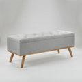 A rectangular upholstered storage bench with a tufted light gray fabric seat and natural wood legs, shown on a plain light gray studio background.