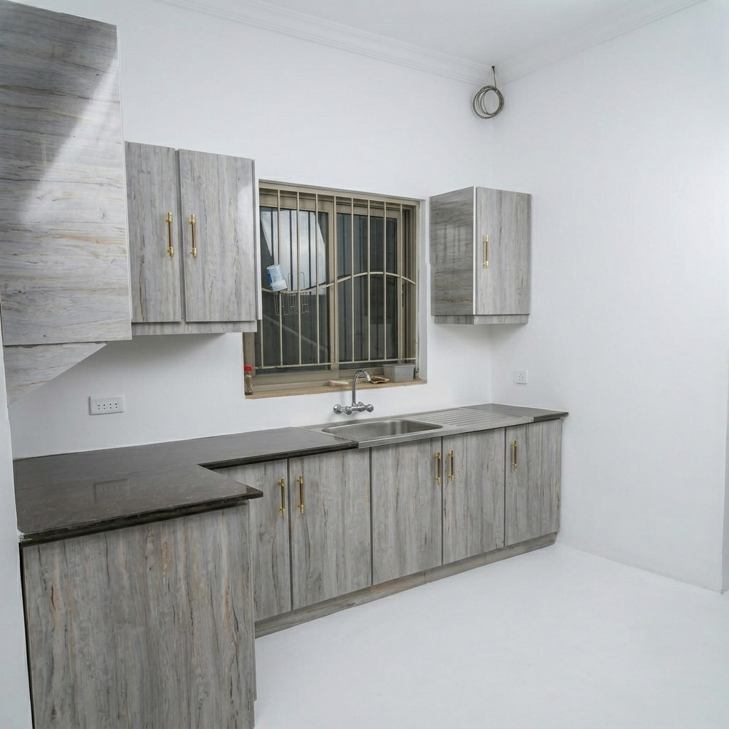 A compact kitchenette setup with gray wood-grain cabinets, matching upper wall cabinets, a dark countertop, a built-in sink, and a barred window set in a white room.