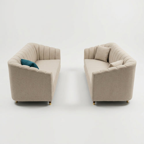 Two modern upholstered armchairs in a light beige fabric with vertical channel tufting, shown side by side on a clean white studio background. One chair includes a teal accent pillow, and both have small gold-toned feet.